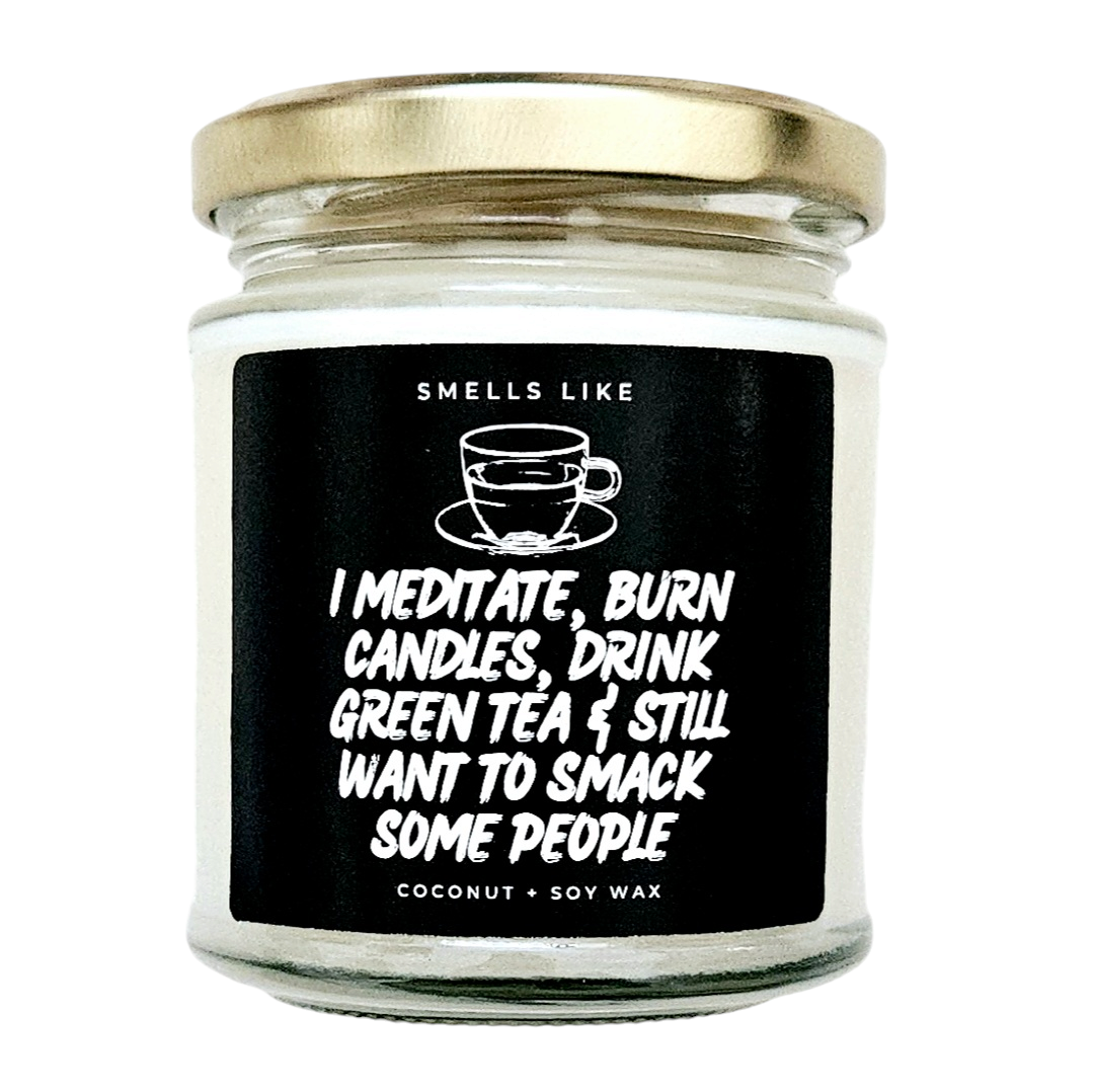 A clear jar candle with a black label that reads 'I Meditate, Burn Candles, Drink Green Tea & Still Want to Smack Some People' along with the scents listed and 'Coconut + Soy Wax'.