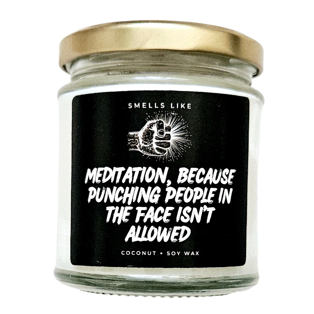 This image features a charming scented candle jar, with a label that reads "Meditation, because punching people in the face isn't allowed". The jar is made of clear glass and is fitted with a shiny gold lid, indicating a stylish yet simple aesthetic.