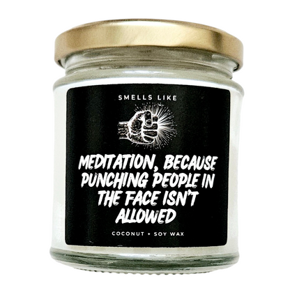 This image features a charming scented candle jar, with a label that reads "Meditation, because punching people in the face isn't allowed". The jar is made of clear glass and is fitted with a shiny gold lid, indicating a stylish yet simple aesthetic.