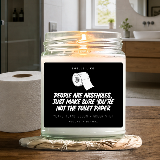 A lit candle in a clear glass jar sits on a wooden bathroom counter, with a roll of toilet paper beside it. The front label is black and white with the text 'SMELLS LIKE PEOPLE ARE ARSEHOLES, JUST MAKE SURE YOU'RE NOT THE TOILET PAPER' and scent text 'YLANG YLANG BLOOM + GREEN STEM' and 'COCONUT + SOY WAX'. By Astrollogy Candles