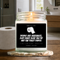 A lit candle in a clear glass jar sits on a wooden bathroom counter, with a roll of toilet paper beside it. The front label is black and white with the text 'SMELLS LIKE PEOPLE ARE ARSEHOLES, JUST MAKE SURE YOU'RE NOT THE TOILET PAPER' and scent text 'YLANG YLANG BLOOM + GREEN STEM' and 'COCONUT + SOY WAX'. By Astrollogy Candles