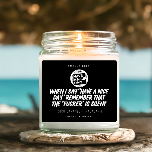 A lit candle in a clear glass jar sits on a woven coaster outdoors near a beach, with a black front label reading “SMELLS LIKE WHEN I SAY ‘HAVE A NICE DAY!’ REMEMBER THAT THE ‘FUCKER' IS SILENT” and scent text “COCO CARAMEL + MACADAMIA.” by Astrollogy Candles