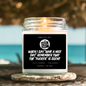 A lit candle in a clear glass jar sits on a woven coaster outdoors near a beach, with a black front label reading “SMELLS LIKE WHEN I SAY ‘HAVE A NICE DAY!’ REMEMBER THAT THE ‘FUCKER' IS SILENT” and scent text “COCO CARAMEL + MACADAMIA.” by Astrollogy Candles
